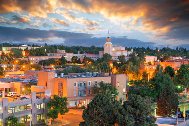 Downtown Santa Fe skyline at sunset with adobe-style buildings, trees, and mountains in the distance.